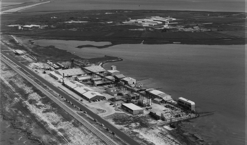 Luftfoto fra 1955. Cheminova begyndte sin produktion på Harboøre Tange i 1953. I baggrunden kunne man allerede i 1955 se de første bygninger til det, der i dag er Cheminovas/FMC Site Rønlands fabriksområde. Foto: Sylvest Jensen Luftfoto, Det Kgl. Bibliotek 